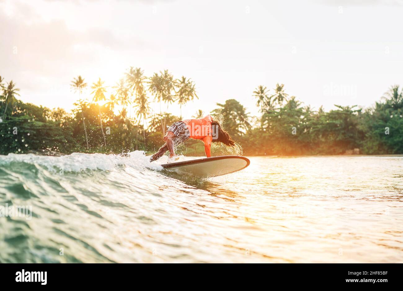 Schwarzer langhaariger Teenager-Junge, der von einem langen Surfbrett reitet und springt. Er fing eine Welle in einer Bucht des indischen Ozeans mit magischem Sonnenuntergang Hintergrund. Extreme Wa Stockfoto