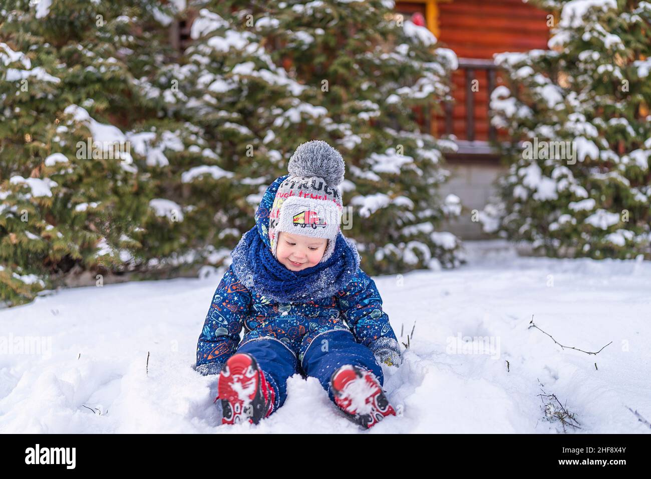 Der kleine Junge sitzt im Schnee in der Nähe des Hauses im verschneiten Waldwinter Stockfoto