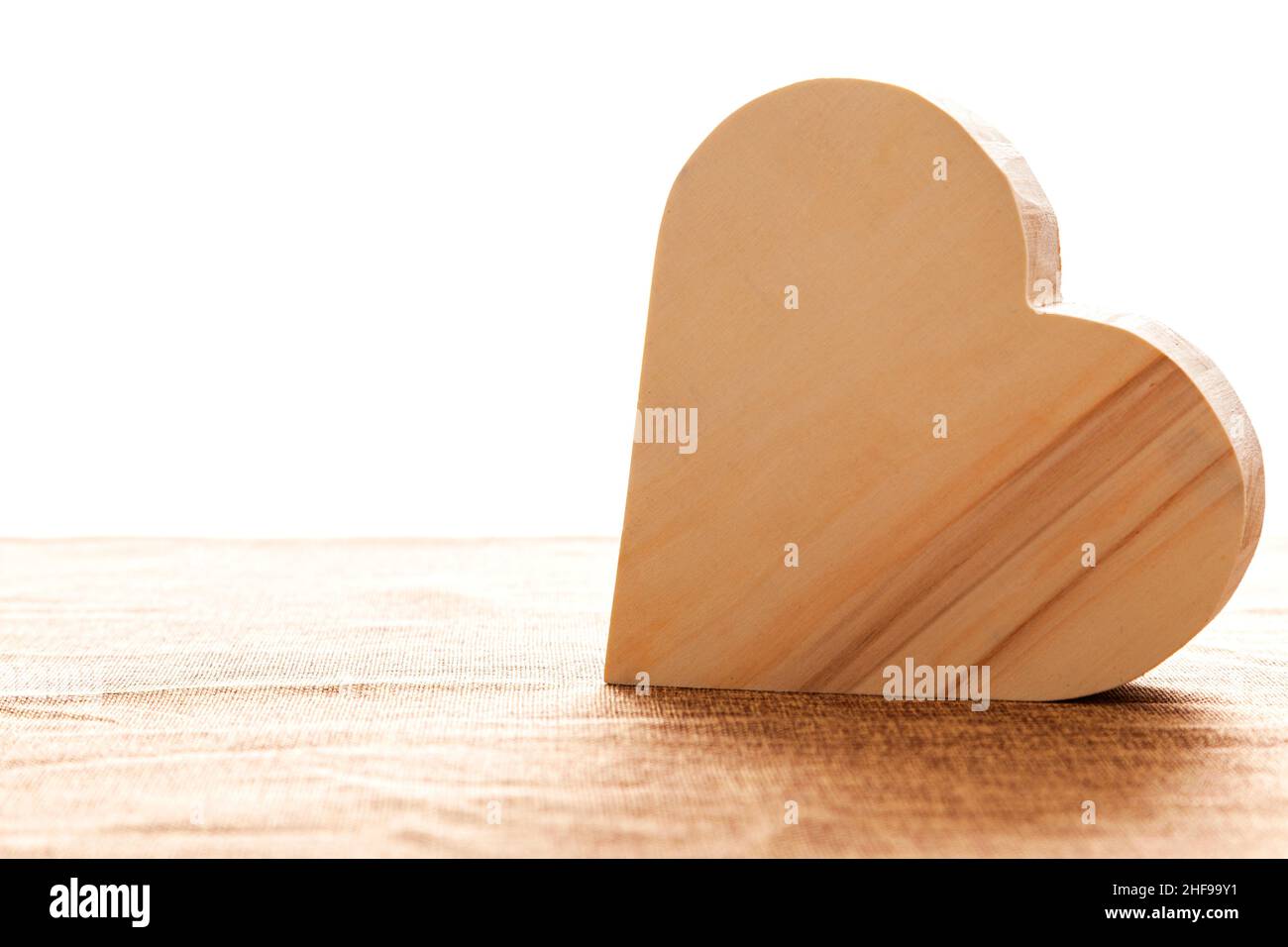 Auf einer Sackleinen-Oberfläche befindet sich ein großes Holzherz. Viel weißes Licht dringt in den Hintergrund ein und wirft einen Schatten vor die Form. Stockfoto