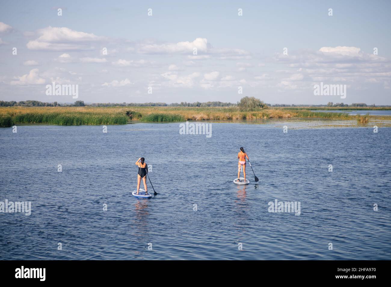 Zwei Frauen mittleren Alters supboarden inmitten eines kleinen blauen Sees, der mit Wellen bedeckt ist, mit Pfützen, die in Badeanzügen in eine Richtung schauen Stockfoto