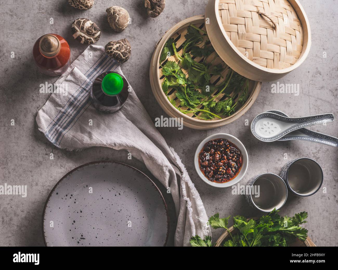 Asiatische Lebensmittel auf grauem Beton: Shiitake-Pilze, dampfender Korb, Hoisin-Sauce, Koriander und würziges Dip, leerer Teller und traditionelles Geschirr. Essen BA Stockfoto