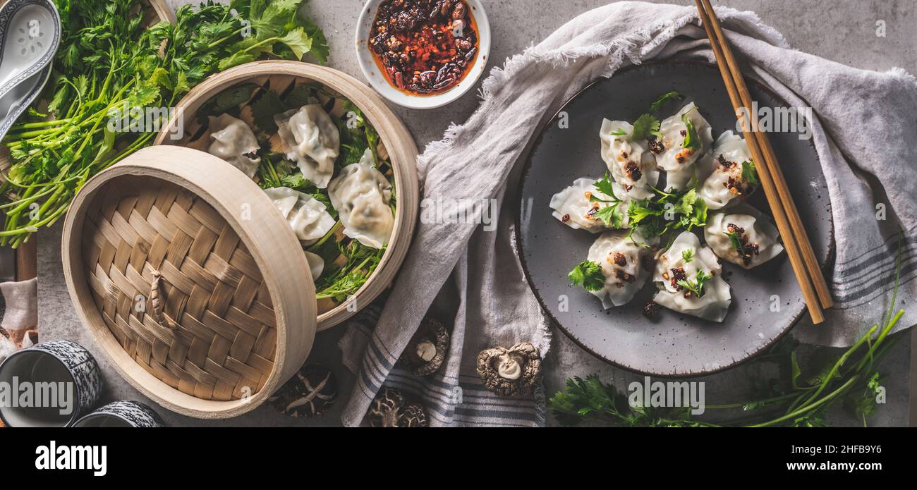 Leckere Knödel im Dampfkorb und auf dem Teller mit Essstäbchen, Kräutern und Dips. Asiatische Küche. Draufsicht. Stockfoto