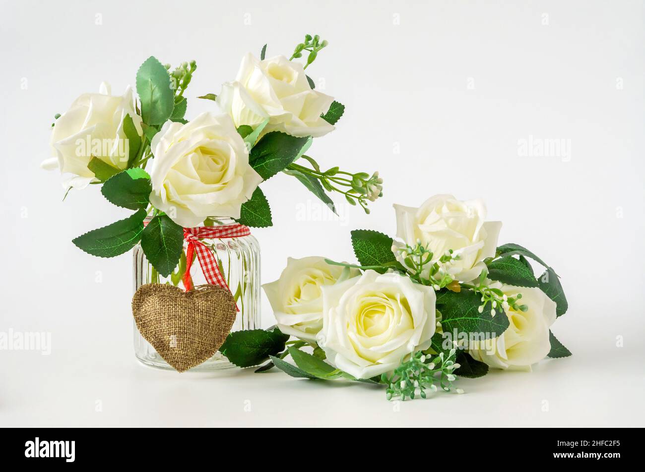 Stillleben, florale Komposition auf weißem Hintergrund, Rosen in einem Glas mit Schleife und Herz Stockfoto