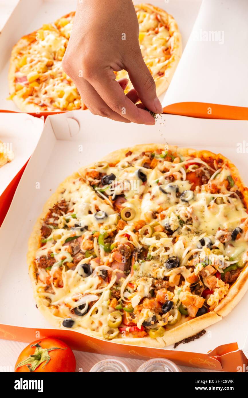 Die Hand eines Menschen legt den Olegano auf eine verzehrfertige Pizza, die Zubereitung von Speisen im Studio, Fast Food mit verschiedenen Zutaten und Verpackungen Stockfoto