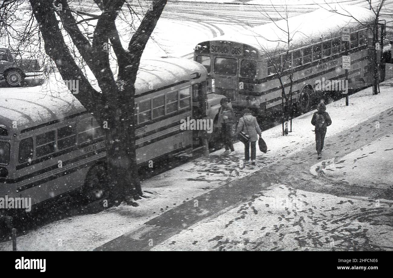 1970s, historisch, winterlich und inmitten von eiskalten Schnee, warten draußen auf der Straße zwei Schulbusse für Schüler, Harlan, Iowa, USA. Die Bereitstellung von Transport für Schulkinder ist in Amerika ein riesiges Unterfangen. Ungewöhnlich in einem kapitalistisch angetriebenen Land war es traditionell ein sozialisiertes System, bei dem lokale öffentliche Einrichtungen kostenlose Schultransporte für Kinder anbieten - manchmal abhängig davon, wie weit sie von den Schulen leben - aus lokalen Steuergeldern und Bundesmitteln. Der Schulbus ist ein ikonischer Teil der amerikanischen Kultur Stockfoto