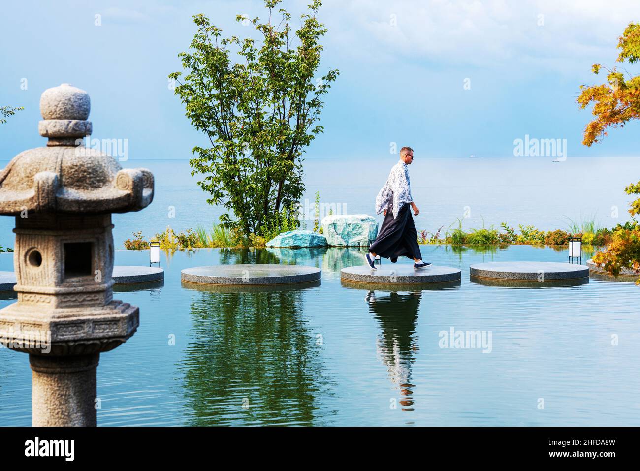 Der Arbeitgeber in traditioneller japanischer Kleidung des japanischen Gartens „Six Senses“ spaziert auf dem Wasser (Teich) auf den runden Steinen des Fußweges. Stockfoto