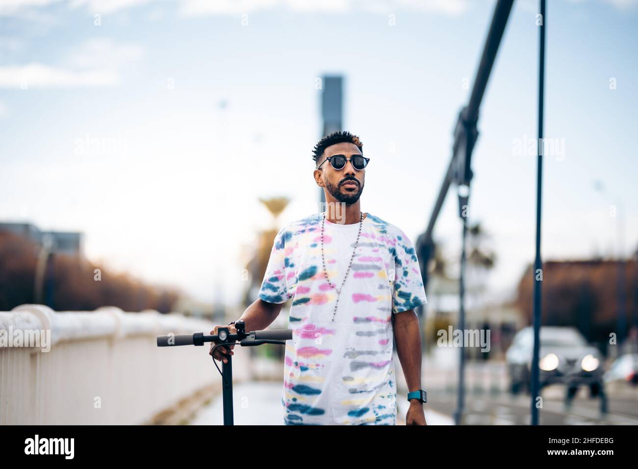 Junger schwarzer Mann im bunten T-Shirt, der mit einem Elektroroller in der Hand spaziert Stockfoto