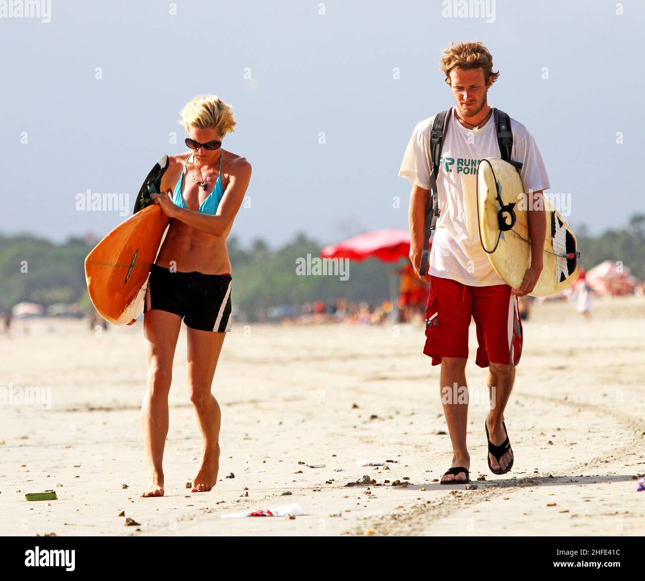 Ein junges blondes Paar, das am Strand entlang läuft, während es seine Surfbretter am Kuta Beach in Bali in Indonesien trägt. Stockfoto