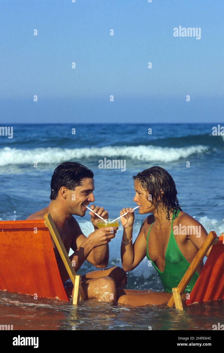 Junge dunkle Haare Paar sitzen Gesicht sehen einander sitzen auf langen Stuhl klares Meerwasser Sommerferien trinken gleichen coktail Stockfoto