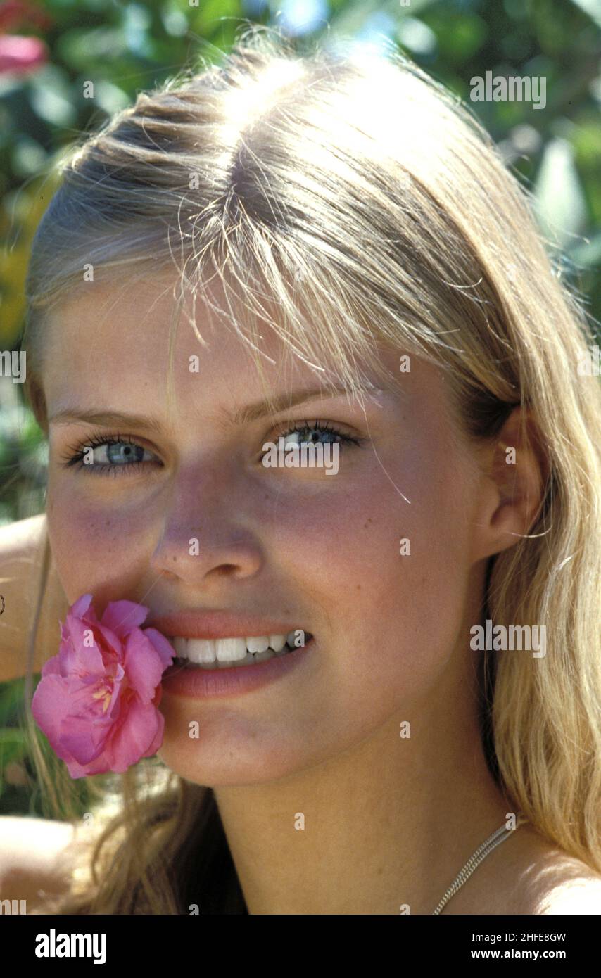 Blonde Haare ziemlich junge Womafront Ansicht natürliches Porträt Vorderansicht Kamera blau lächelnd Blume Lippen grün natürliches Laub Hintergrund Stockfoto