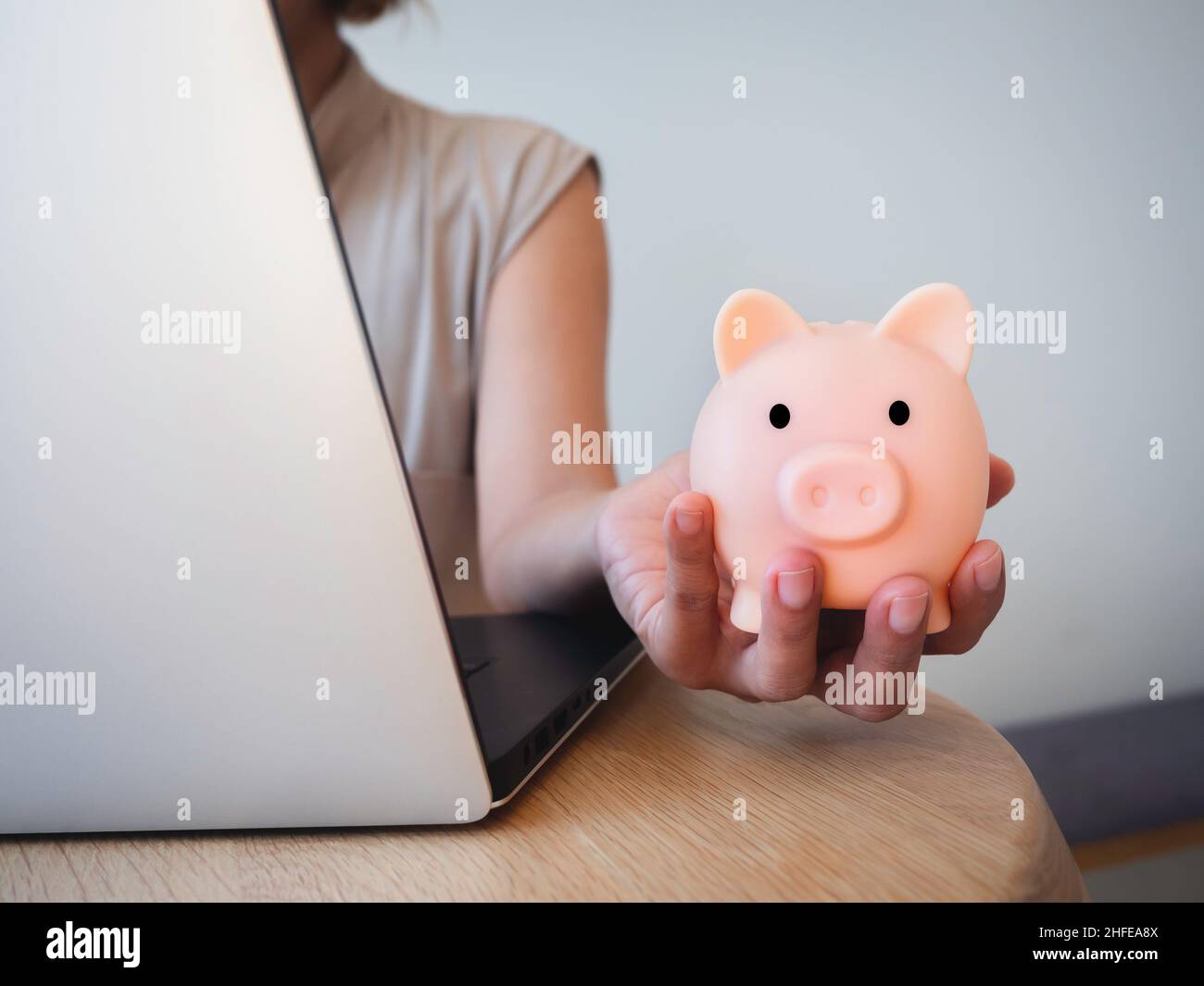 Geldbox zur Hand. Die Hand hält das pinke Sparschwein, während man den Laptop auf dem Schreibtisch benutzt. Geld sparen Reichtum und Finanzkonzept, Geschäft, online b Stockfoto