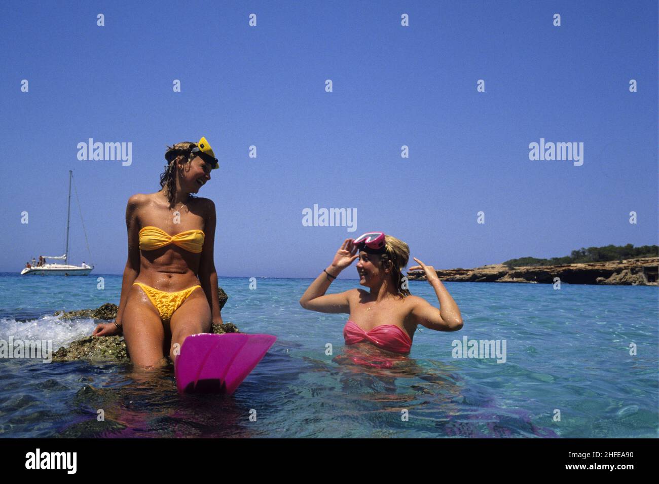 2 hübsche blonde Haare junge Frau Vorderansicht Scuba Diving Maske sitzen auf Rock blau siehe Hintergrund Stockfoto