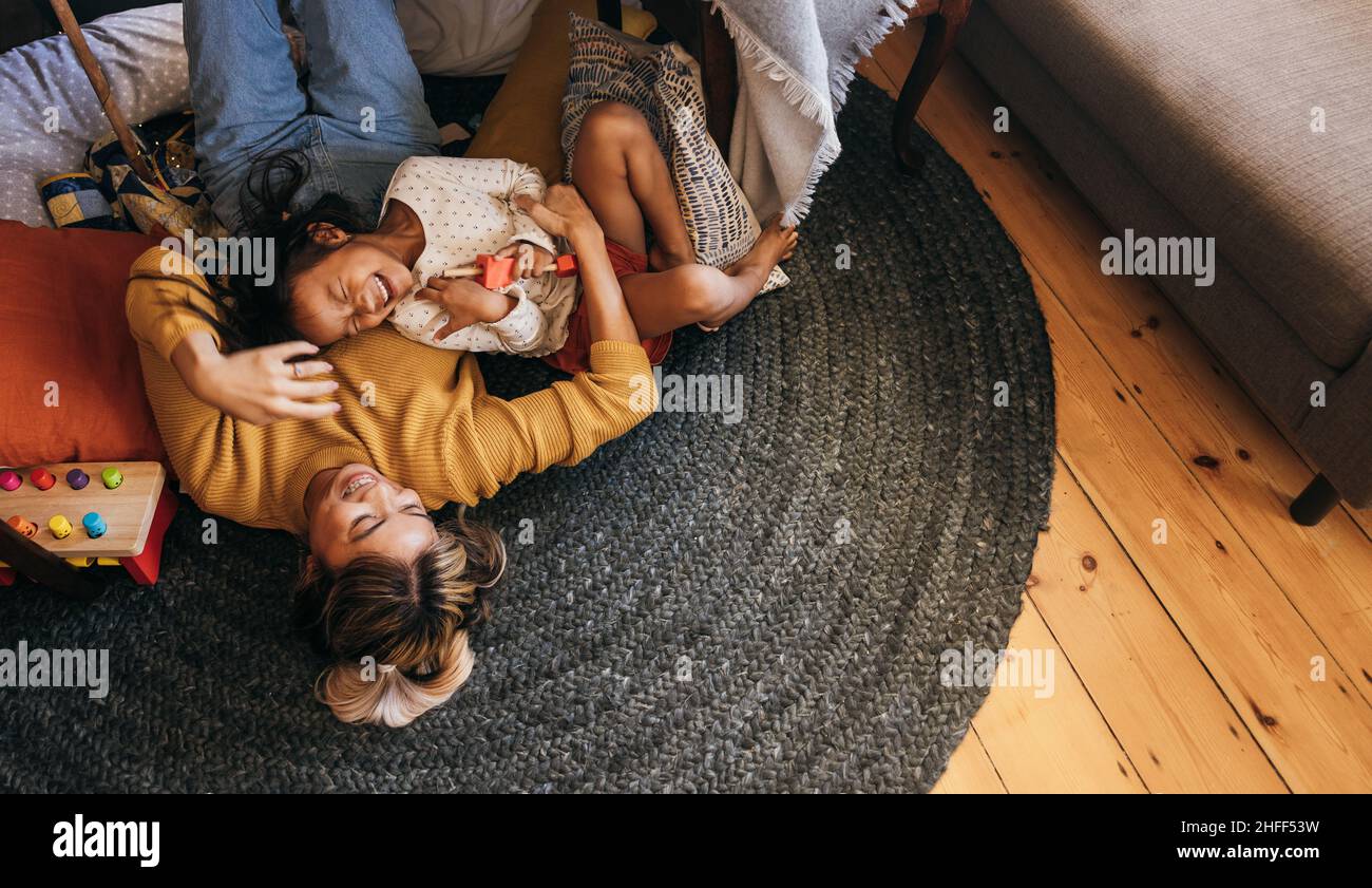 Blick von oben auf eine Mutter und ihre Tochter, die zu Hause fröhlich lachen. Fröhliche Mutter und Tochter liegen auf dem Boden in ihrem Spielbereich. Mutter und da Stockfoto