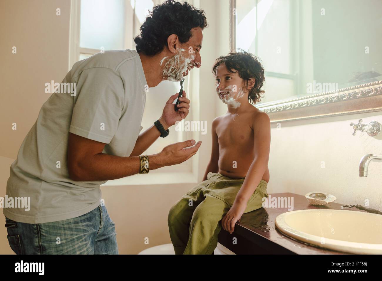 Fröhlicher Vater, der seinem Sohn zeigt, wie man sich zu Hause mit Rasiercreme rasieren kann. Glücklicher Vater, der seinem Sohn im Badezimmer seine Rasierkünste beibrachte. Vater und Stockfoto