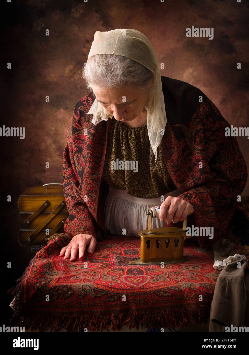 Alte Frau im viktorianischen Kleid, die mit einem antiken Holzkohleisen auf einem Brett arbeitet Stockfoto