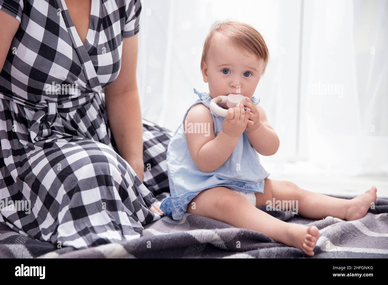 Das kleine Mädchen beißt einen Beißring aus Holz und ihre Mutter sitzt nahe bei ihr Stockfoto