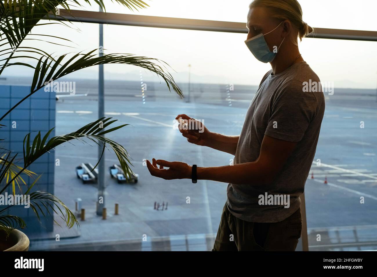Mann, der am Flughafen oder an öffentlichen Orten Desinfektionsmittel auf die Hände sprüht. Hintergrundbeleuchtung vom Fenster, Silhouette. Mann in Gesichtsmaske, Flug und Covid Sicherheit Stockfoto