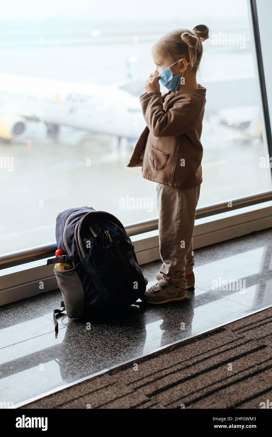 Kind mit Rucksack, das durch das Flughafenfenster auf das Flugzeug schaut. Sichere Reise mit Kindern während Covid Ausbruch in Gesichtsmaske. Kind Passagier bei der Ankunft Stockfoto