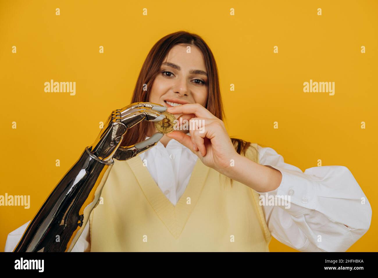 Lady mit modernen glänzenden Metall bionischen Arm hält große goldene Bitcoin posiert für die Kamera auf gelbem Hintergrund extreme Nahsicht Stockfoto