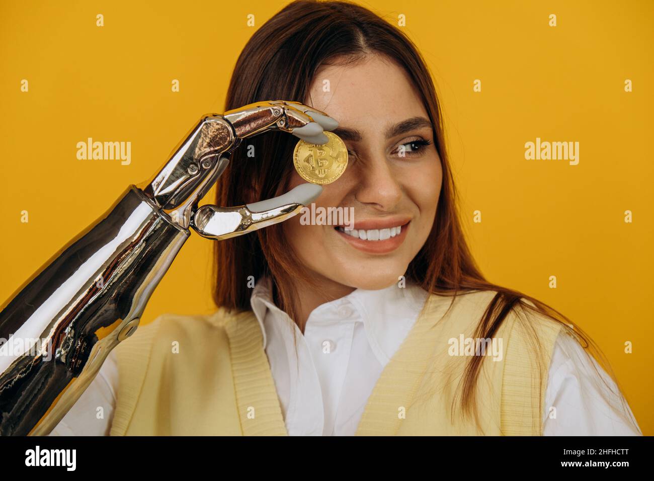 Lady mit modernen glänzenden Metall bionischen Arm hält große goldene Bitcoin posiert für die Kamera auf gelbem Hintergrund extreme Nahsicht Stockfoto