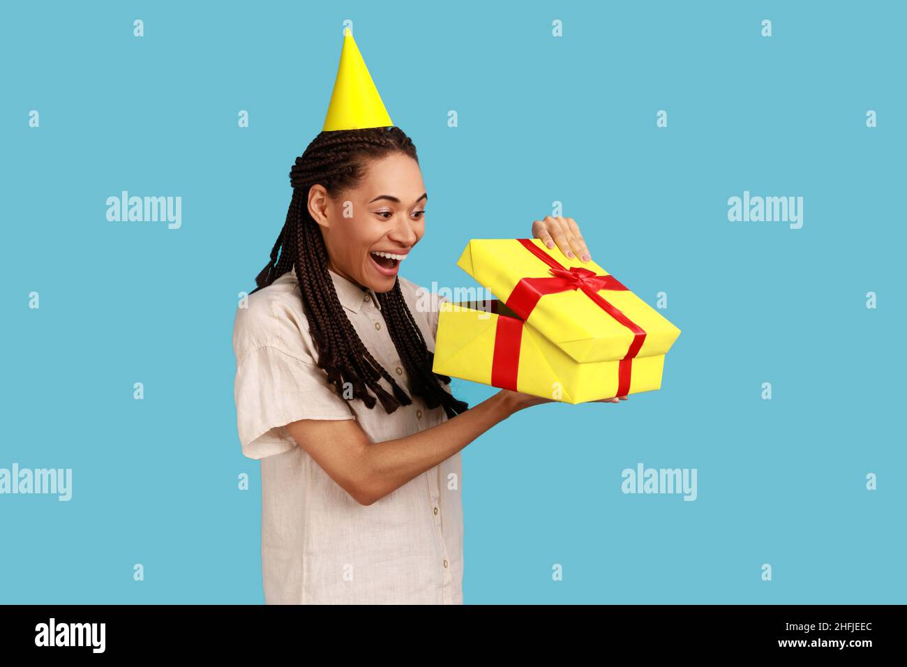 Porträt einer staunenden Frau in gelbem Partykegel, die in die Geschenkbox schaut, das Geschenk öffnet und voller Aufregung in das Innere blickt, das weißes Hemd trägt. Innenaufnahme des Studios isoliert auf blauem Hintergrund. Stockfoto