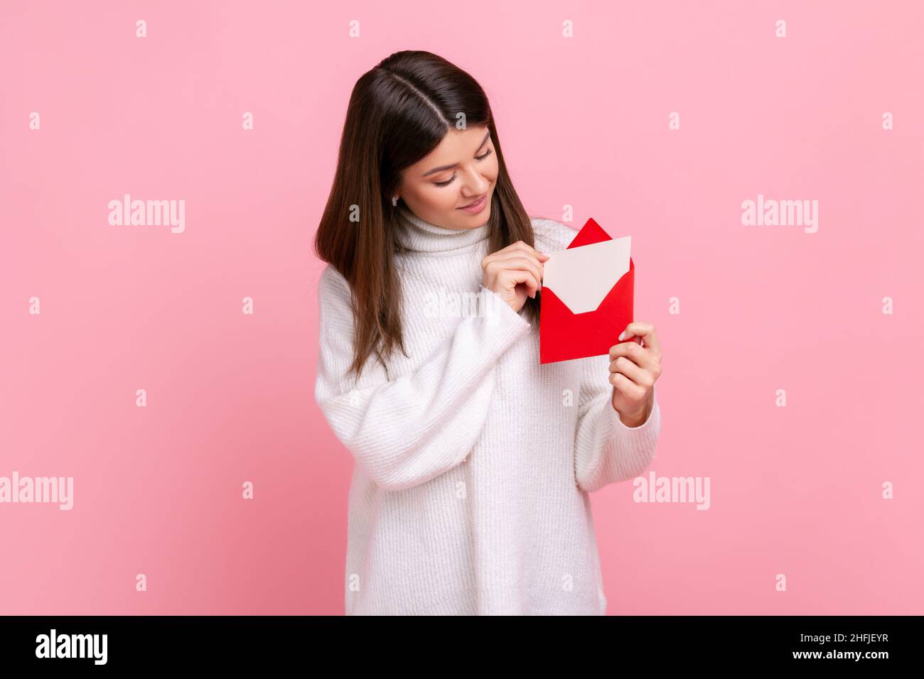 Porträt einer glücklichen Frau mit rotem romantischen Umschlag und Blick auf Brief mit Lächeln und Interesse, tragen weißen lässigen Stil Pullover. Innenaufnahme des Studios isoliert auf rosa Hintergrund. Stockfoto