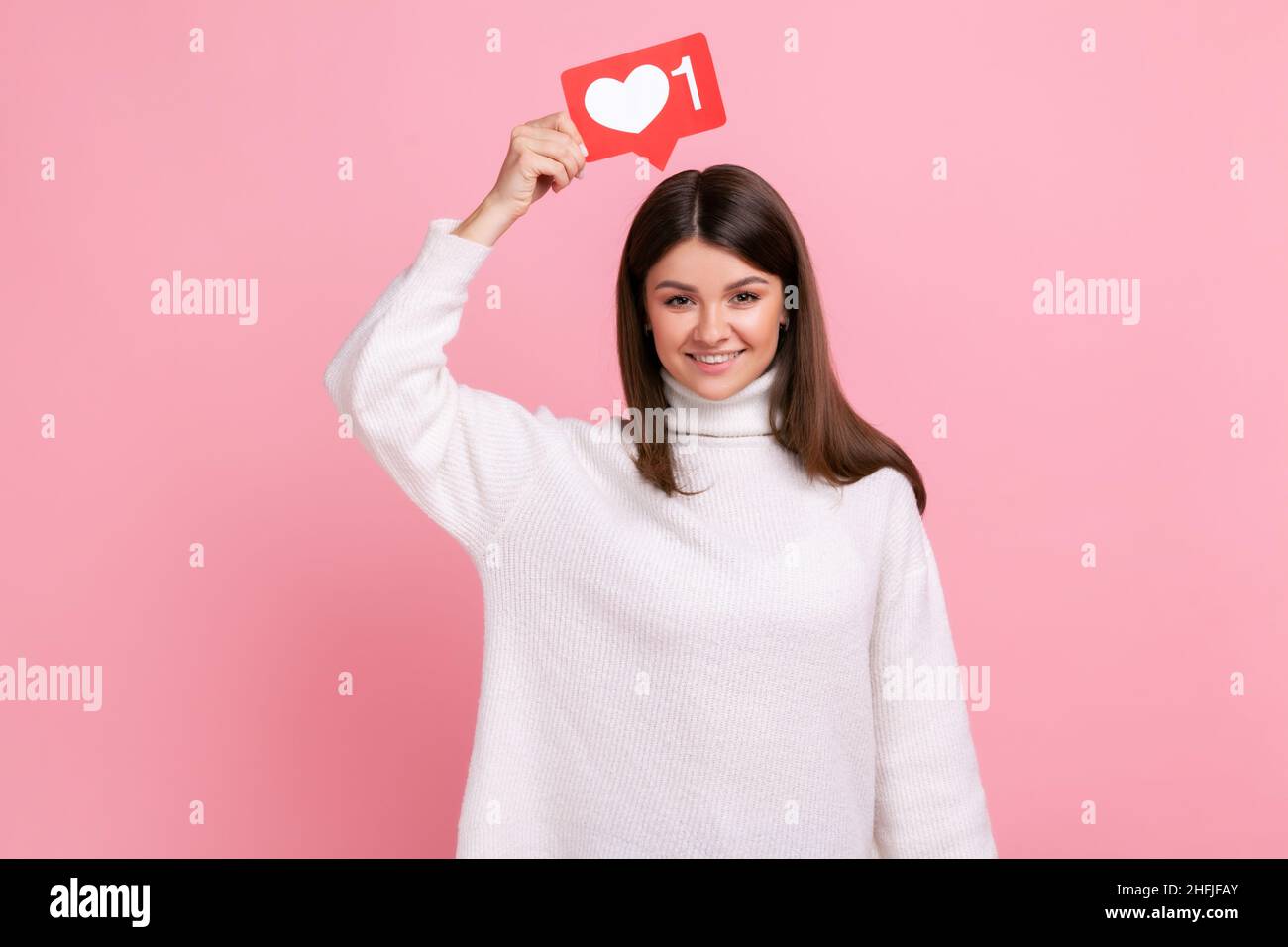 Lächelnde Frau, die Herz-Ikone über den Kopf hält und empfiehlt, dem Blog mit interessanten Inhalten zu folgen und einen weißen Pullover im lässigen Stil zu tragen. Innenaufnahme des Studios isoliert auf rosa Hintergrund. Stockfoto