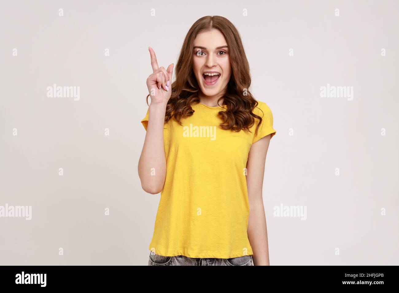 Porträt einer glücklichen inspirierten Frau jungen Alters in gelben Casual T-Shirt mit Finger heben und geniale Idee, die von plötzlichen intelligenten Gedanken erstaunt. Innenaufnahme des Studios isoliert auf grauem Hintergrund. Stockfoto