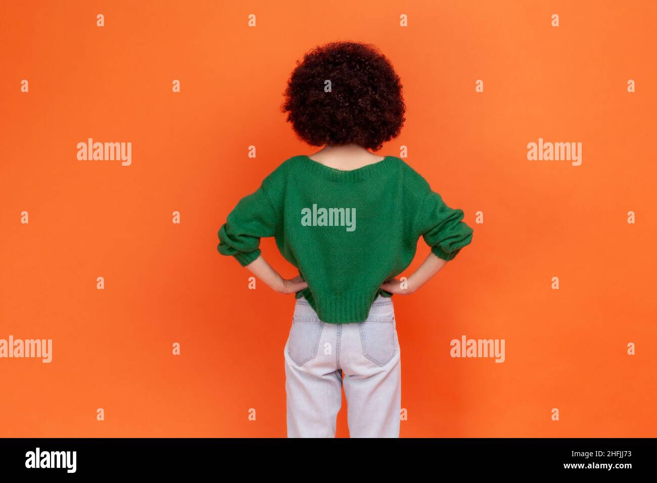 Rückansicht Porträt einer Frau mit Afro-Frisur trägt einen grünen Pullover im lässigen Stil, der nach hinten posiert und die Hände auf den Hüften hält. Innenaufnahme des Studios isoliert auf orangefarbenem Hintergrund. Stockfoto