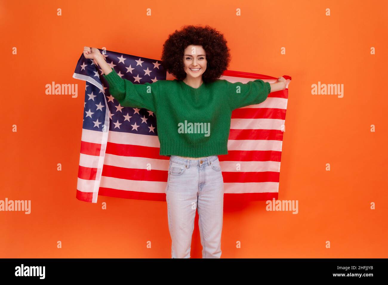 Glücklich positive Frau mit Afro-Frisur trägt grünen lässigen Stil Pullover stehen mit erhobenen Armen, hält die USA-Flagge, feiert nationalen Feiertag. Innenaufnahme des Studios isoliert auf orangefarbenem Hintergrund. Stockfoto