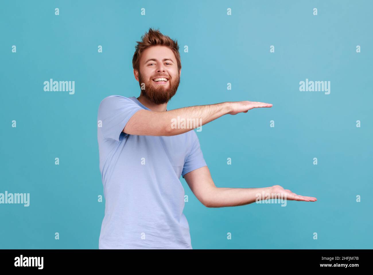 Porträt eines jungen Erwachsenen mit positivem optimistischem Bart, der einen Werbebereich präsentiert und mit einem Lächeln und Optimismus die Kamera anschaut. Innenaufnahme des Studios isoliert auf blauem Hintergrund. Stockfoto