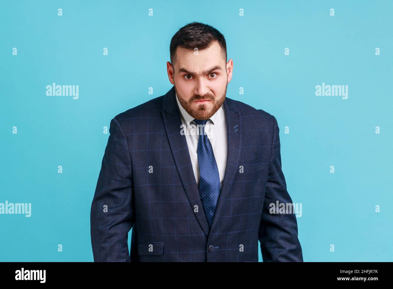 Wütender, gutaussehender Geschäftsmann, der einen Anzug im offiziellen Stil trägt und die Kamera anschaut, mit aggressivem Gesichtsausdruck, der negativ, Hass ausdrückt. Innenaufnahme des Studios isoliert auf blauem Hintergrund. Stockfoto