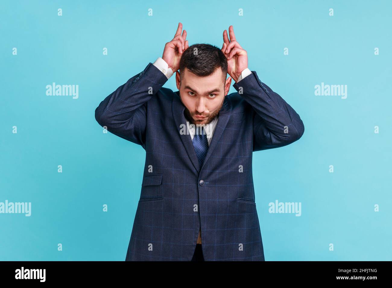 Aggressiver Schläger mit Bart und offiziellem Anzug, der die Finger über dem Kopf hält und Hörner zeigt, arrogant und stur, angriffbereit. Innenaufnahme des Studios isoliert auf blauem Hintergrund. Stockfoto