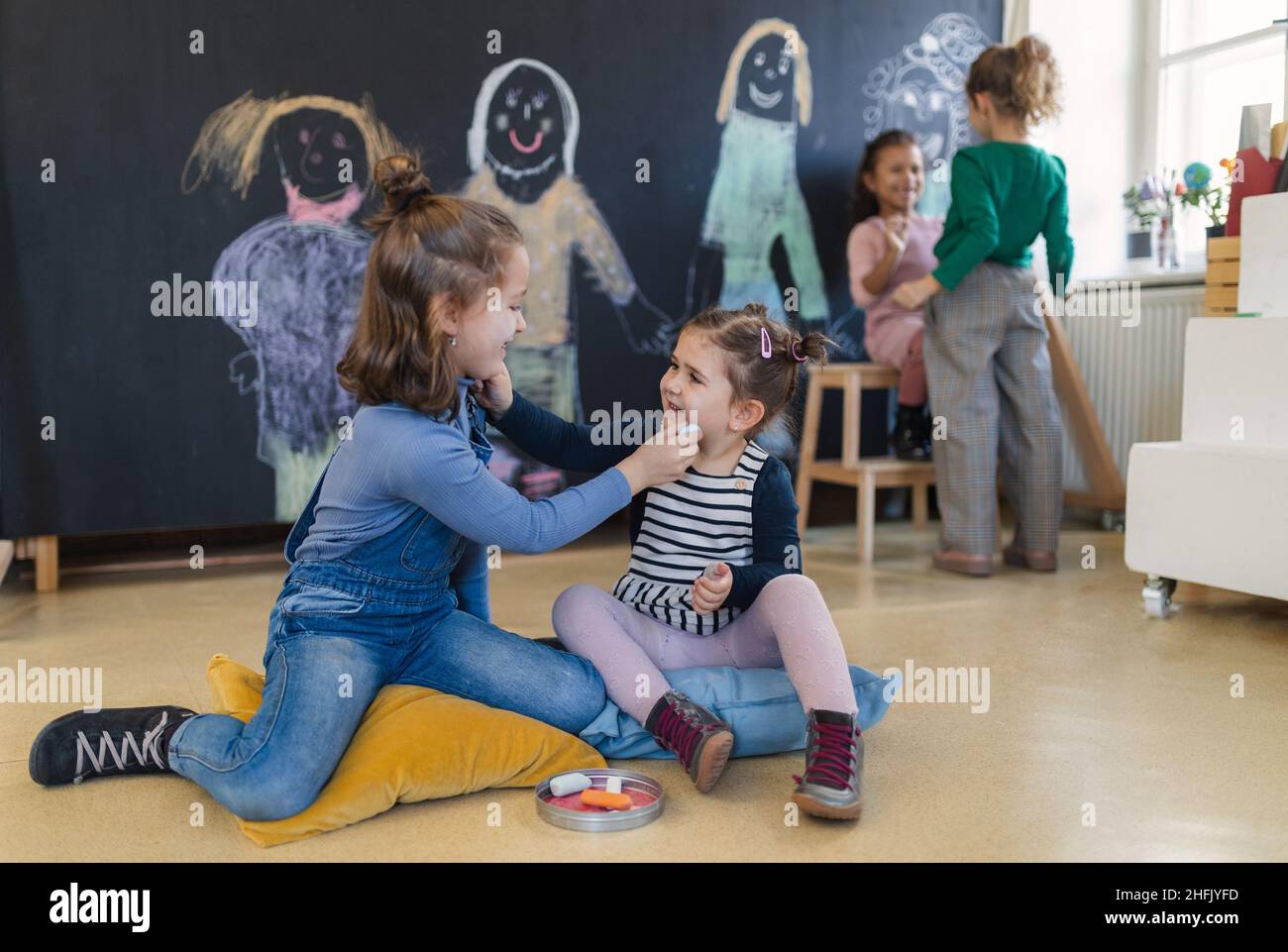 Gruppe kleiner Mädchen, die im Spielzimmer mit Kreiden auf den Wangen des anderen zeichnen. Stockfoto