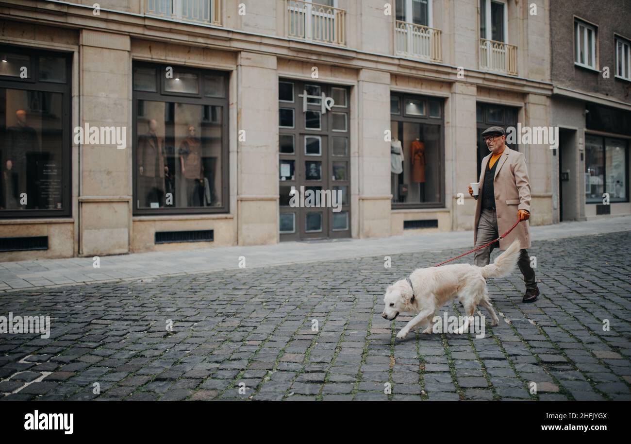 Seitenansicht eines eleganten älteren Mannes mit Kaffee zum Mitnehmen, der im Winter mit seinem Hund in der Stadt unterwegs ist. Stockfoto
