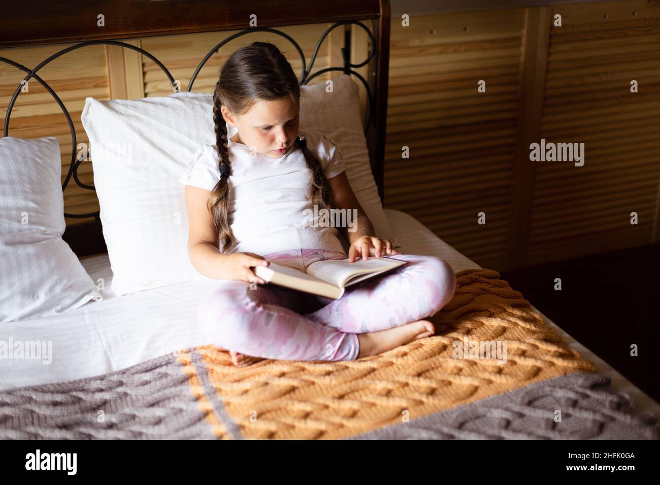 Kleines ernsthaftes Mädchen, das sich das Buch aufmerksam anschaut und auf dem Bett sitzt und Hauskleidung in einem Holzhaus trägt. Freizeitangebote. Selbst Stockfoto