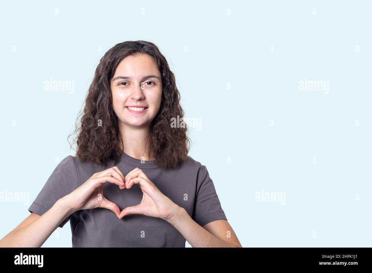 Ein Mädchen mit lockigen Haaren macht Hände in Form eines Herzens auf einem hellblauen Hintergrund. Herzkrankenversicherung, Spende, glückliche Wohltätigkeitsorganisation, worl Stockfoto