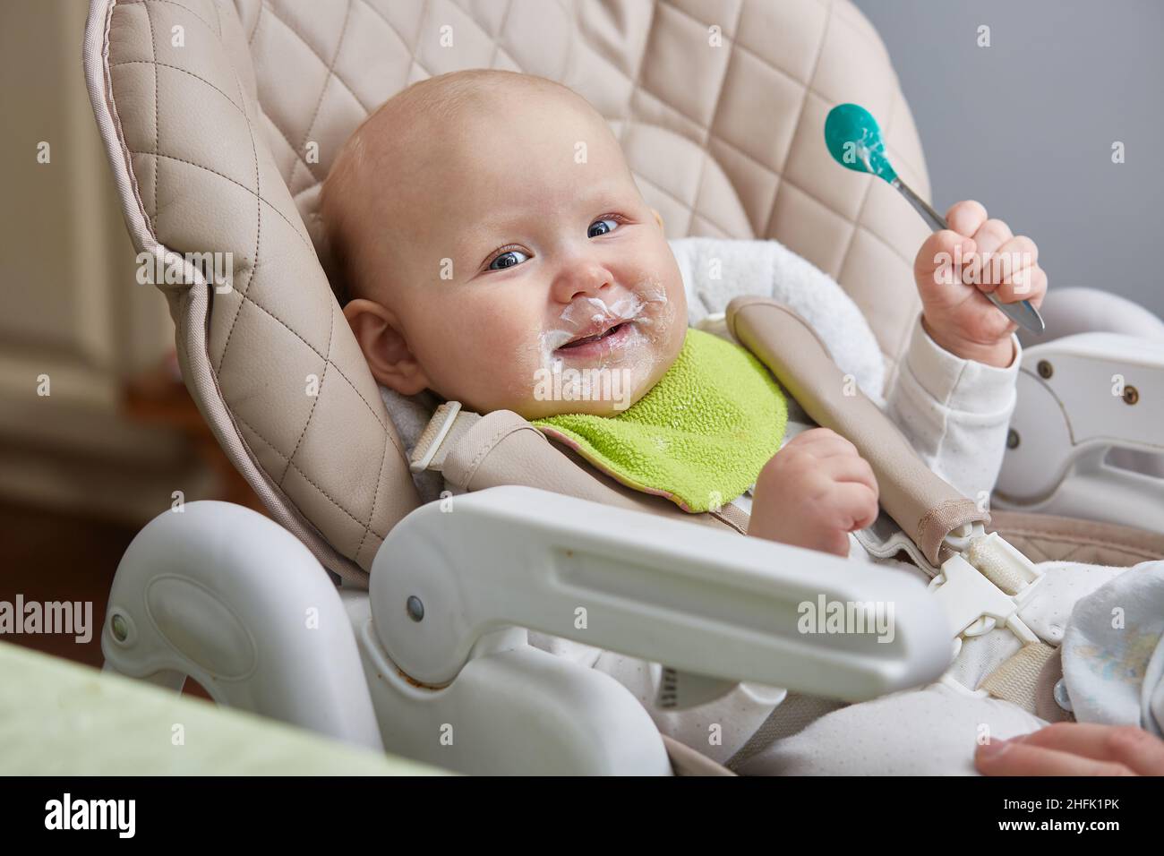 Lächelndes, glückliches Baby mit einem Löffel in den Händen versucht die erste Milchköder Fütterung Stockfoto