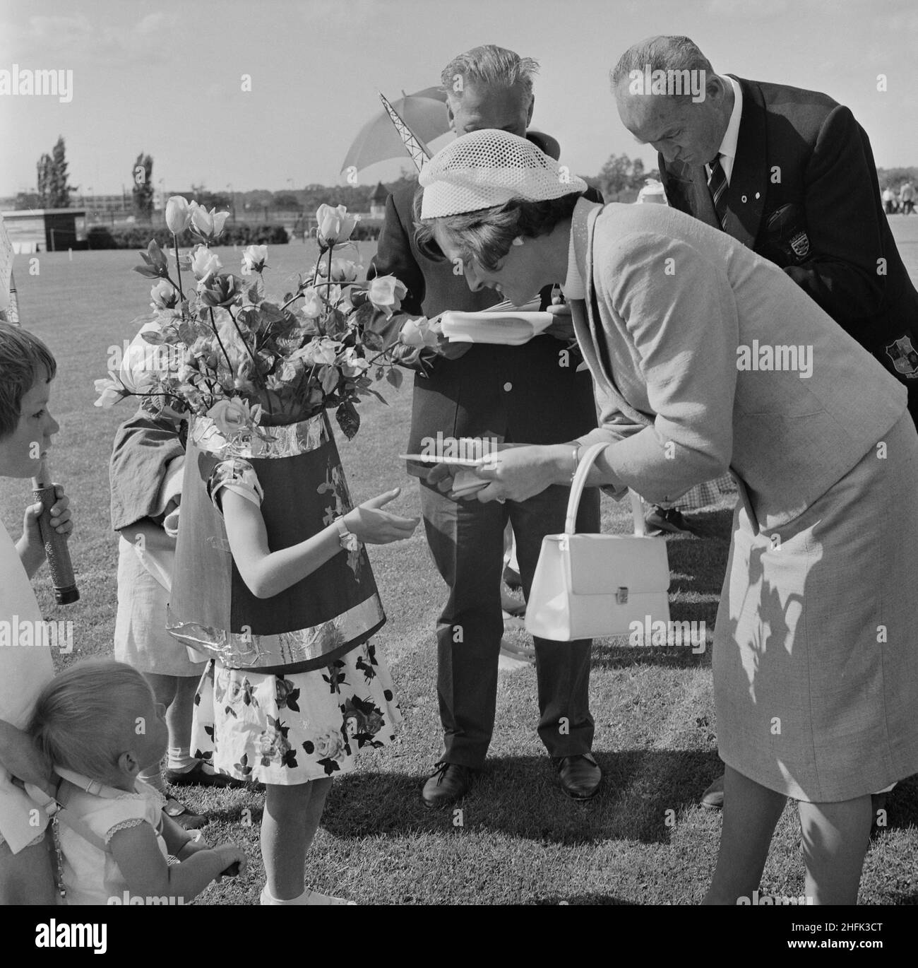 Laing Sports Ground, Rowley Lane, Elstree, Barnett, London, 26/06/1965. Hilda Laing beim jährlichen Laing-Sporttag, bei dem ein kleines Kind, das als Blumenvase gekleidet ist, einen Preis für ein ausgefallenes Kleid überreicht. Im Jahr 1965 wurde Laings jährlicher Sporttag am 26th. Juni auf dem Sportplatz auf der Rowley Lane abgehalten. Neben Fußball und Leichtathletik gab es auch Neuheitenveranstaltungen wie das Sack-Rennen und das Donkey Derby sowie Veranstaltungen für Kinder wie Go-Kart-Rennen, Wettbewerbe für ausgefallene Kleidung und Pony-Fahrten. Stockfoto