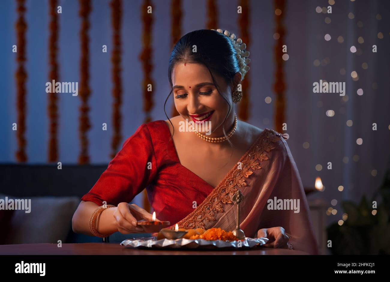 Eine wunderschöne Frau, die ein Diya setzt und lächelt, inmitten von diwali-Dekoration, Lichtern und einem Pooja-Thali. Stockfoto