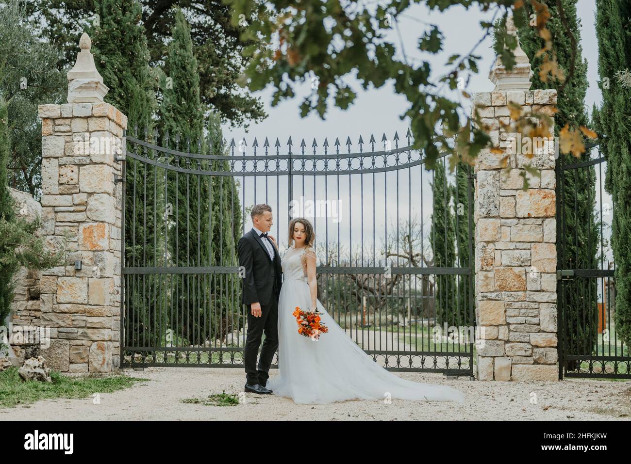 Fröhliches, stilvolles und lächelndes Paar, das am Hochzeitstag in der Toskana, Italien, unterwegs ist. Die Braut und der Bräutigam gehen die Straße entlang durch die Hände. Ein stylischer Junge Stockfoto