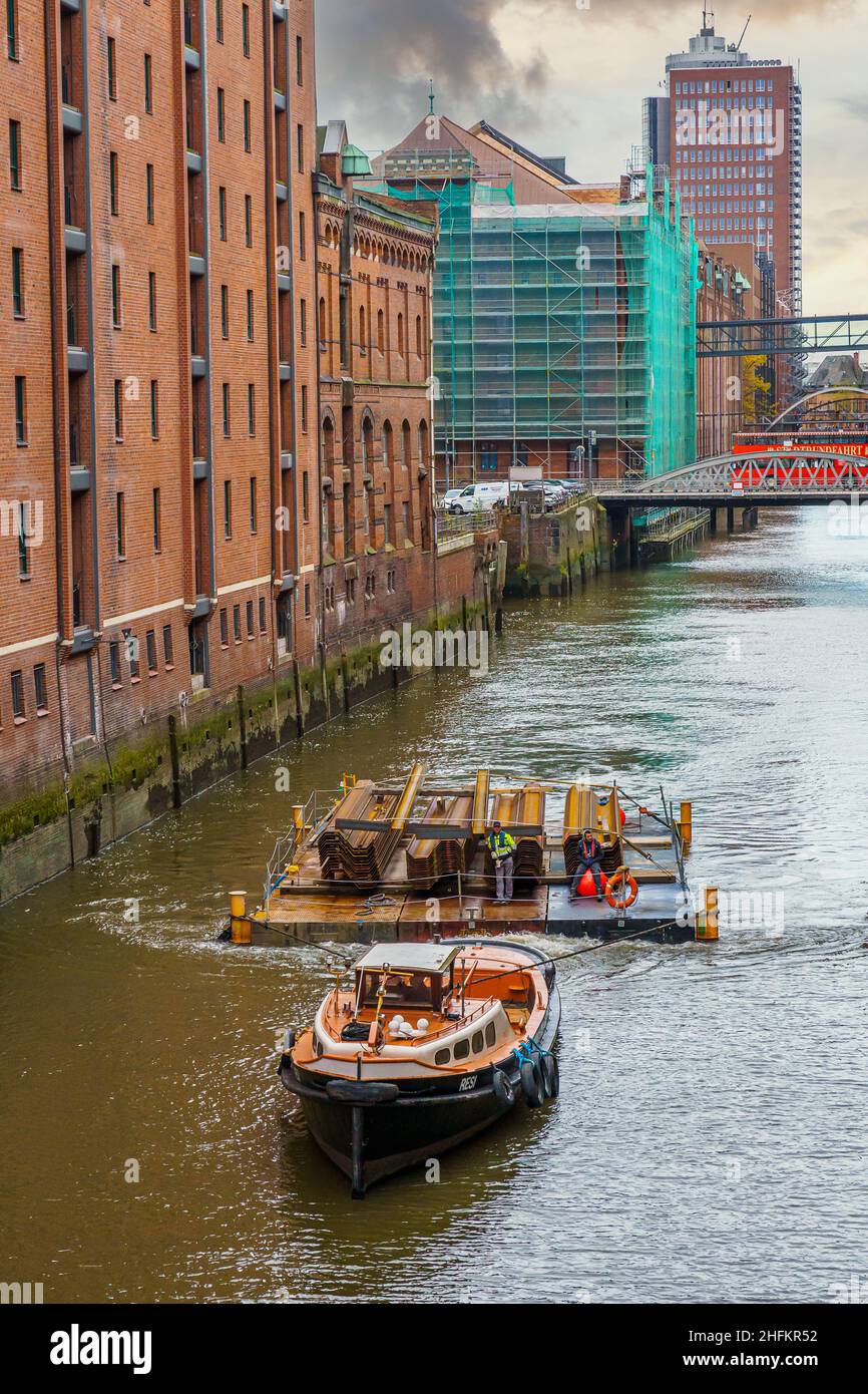 Eine Barkasse zieht auf einem Fleet in der Hamburger Speicherstadt ein Floß oder Ponton mit Stahlplatten und zwei gelangweilten Arbeitern Stockfoto