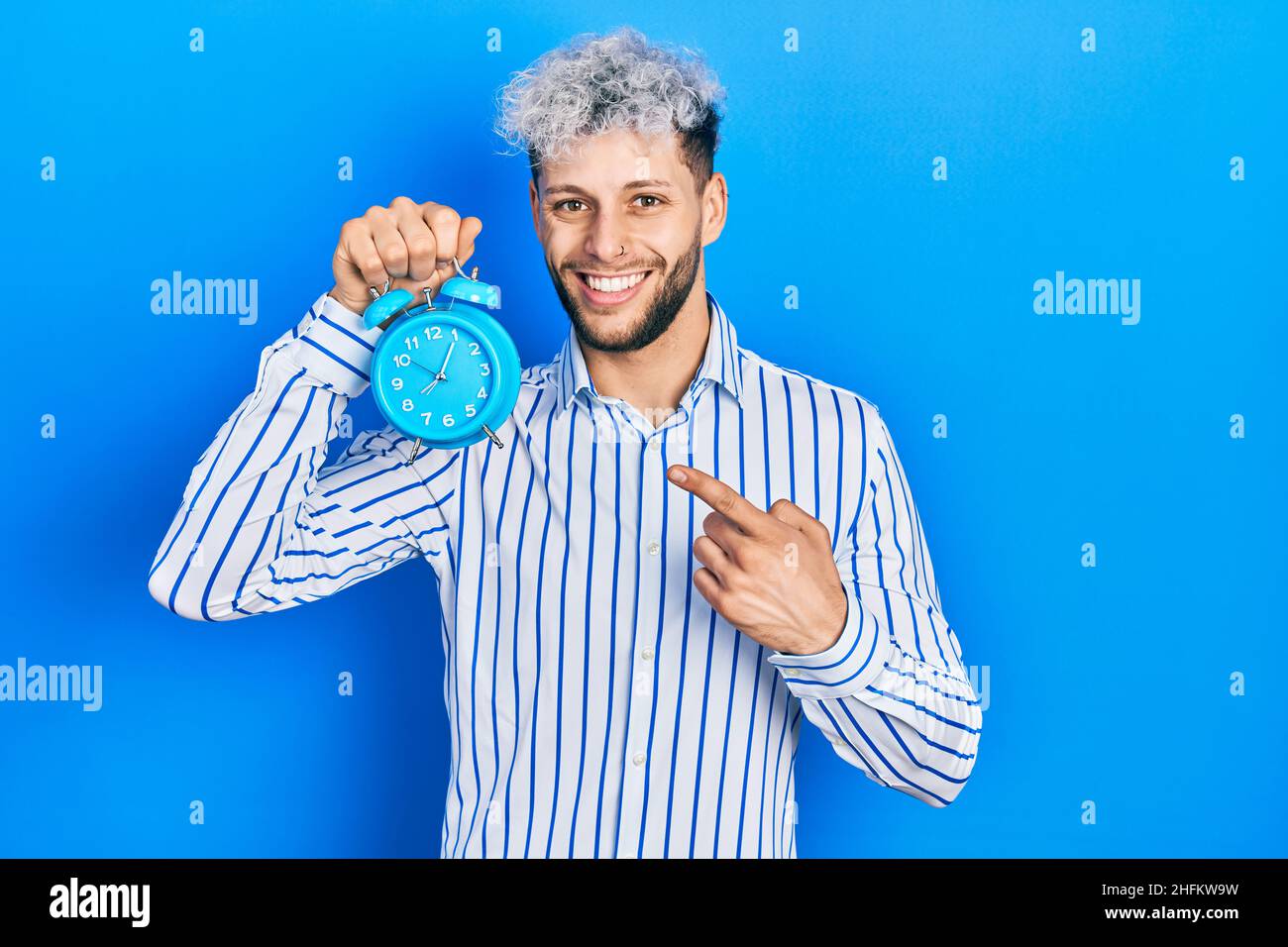 Junger hispanischer Mann mit modernen gefärbten Haaren, die den Wecker lächelnd mit Hand und Finger zeigen Stockfoto