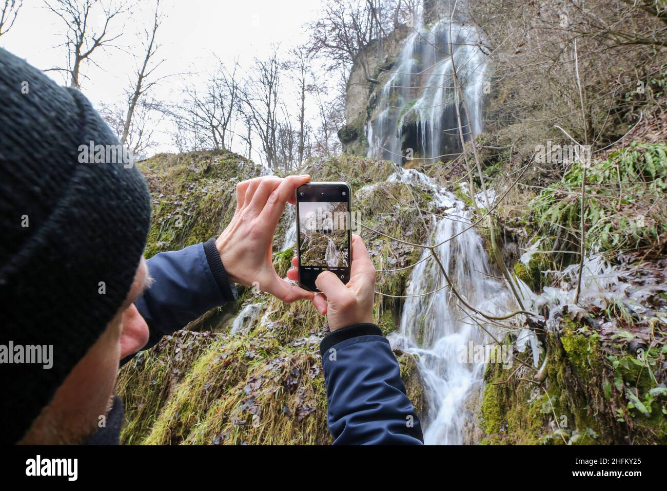 Bad Urach, Deutschland. 16th Januar 2022. Ein Mann fotografiert den Wasserfall bei Bad Urach mit seinem Handy. Der Urach-Wasserfall gehört zu den Sehenswürdigkeiten des UNESCO Global Geopark Schwäbische Alb und des UNESCO-anerkannten Biosphärenreservats Schwäbische Alb. Quelle: Thomas Warnack/dpa/Alamy Live News Stockfoto