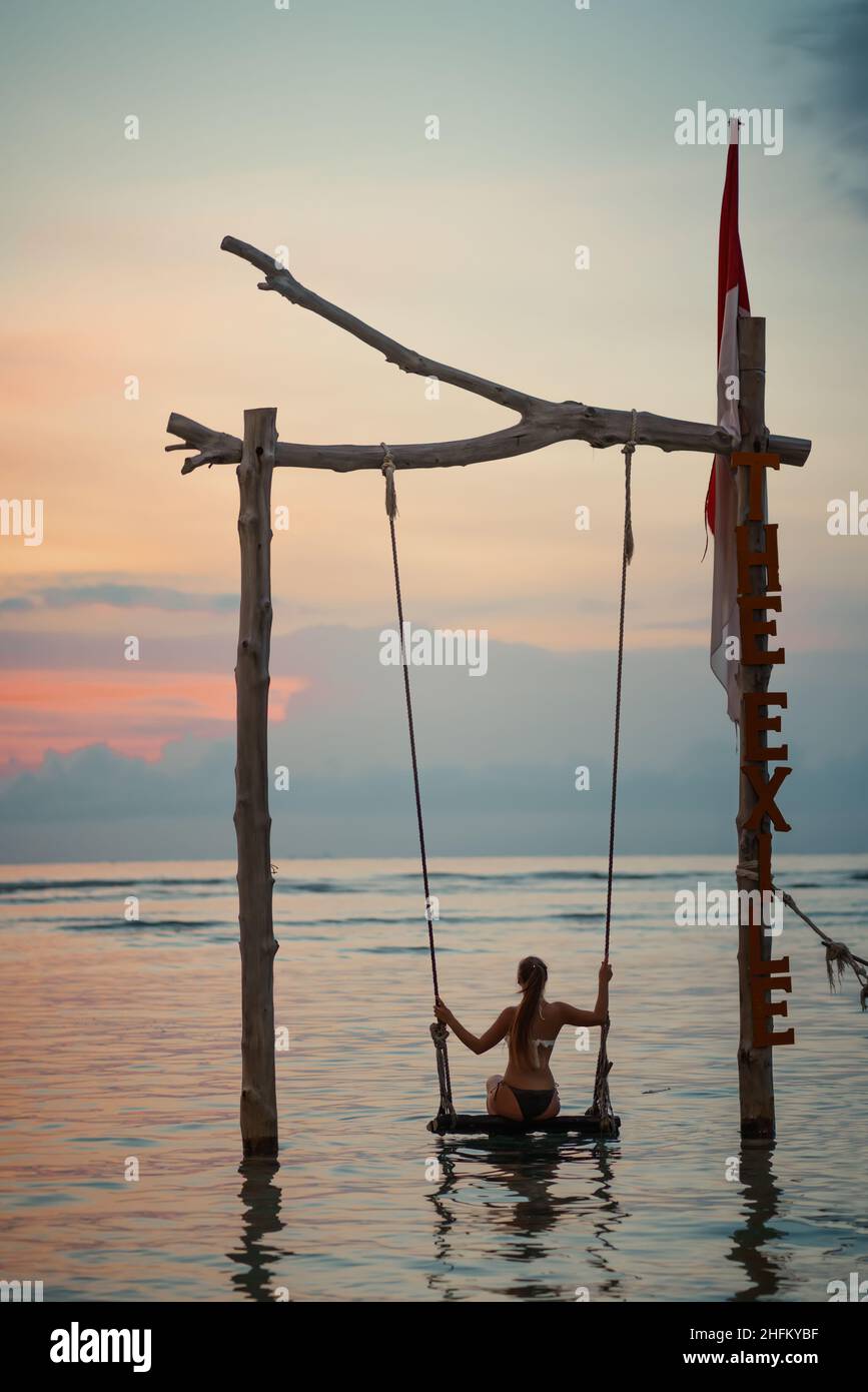 Gebräunte Frau, die auf einer Schaukel im Wasser steht. Sonnenuntergang und Meer im Hintergrund. Relax und Urlaub Konzept. Stockfoto