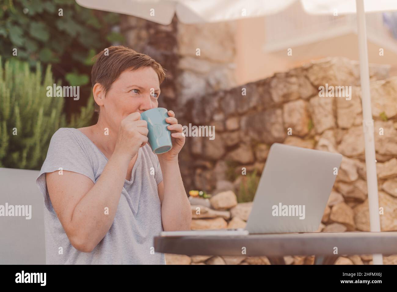 Frau trinkt Kaffee, während sie von der Terrasse auf dem Laptop am Sommermorgen arbeitet Stockfoto