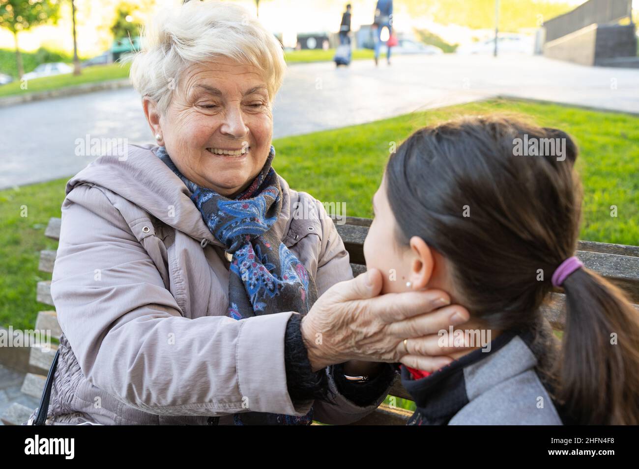 Ältere blonde Frau streichelte Enkelin, die auf der Parkbank sitzt. Großmutter und kleines Mädchen lieben, Zuneigung Konzepte Stockfoto