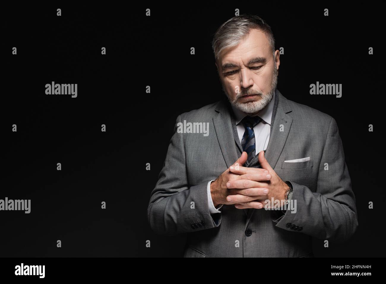 Nachdenklicher älterer Mann in Blazer, der mit zusammengekrallten Händen auf Schwarz isoliert steht Stockfoto