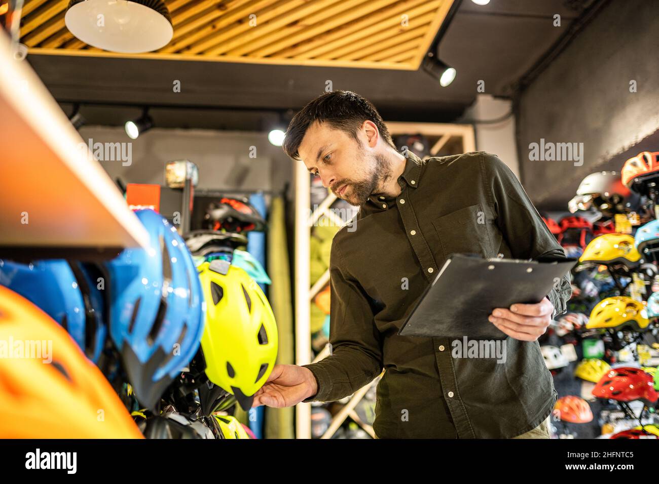 Ein männlicher Bike-Shop-Manager führt in einem Bike-Shop eine Inventarisierung von Sporthelmen durch. Der Besitzer eines Sportladens mit einem Clipboard in den Händen prüft die Stockfoto
