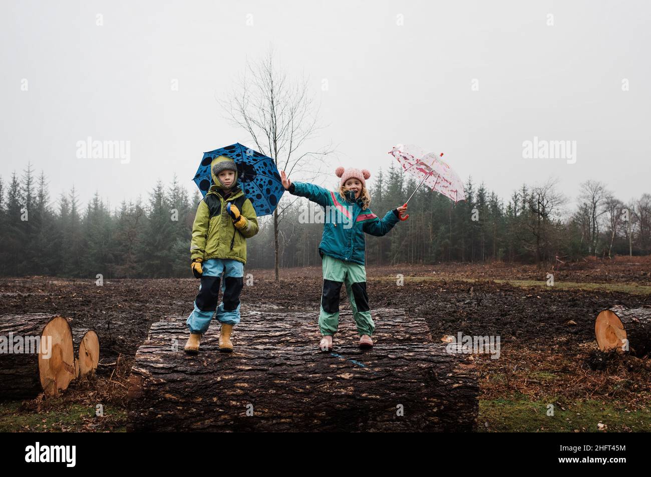 Kinder spielen gerne auf Holzstämmen draußen im Regen mit Regenschirmen Stockfoto